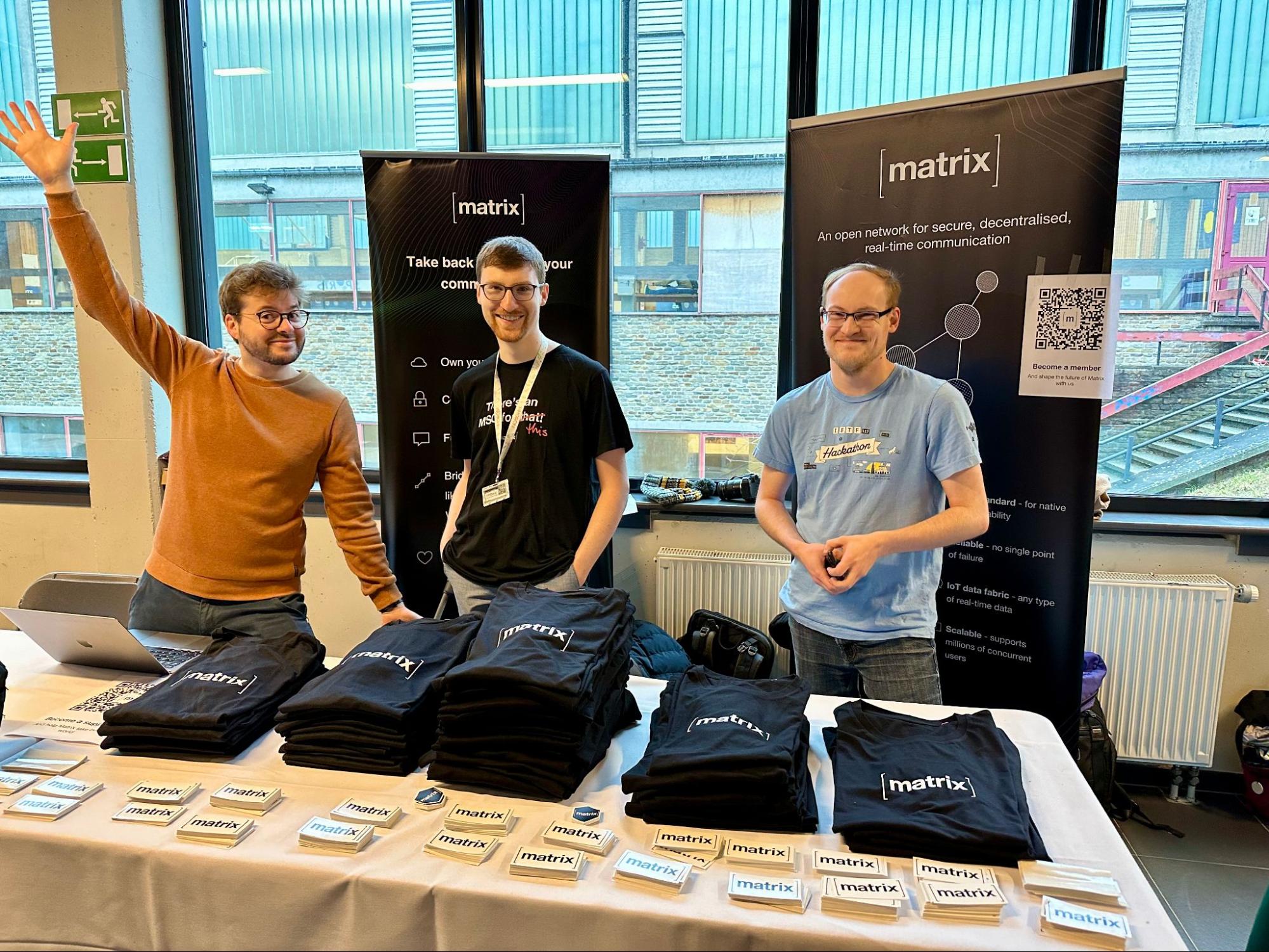 A picture of three male presenting people behind a table, smiling in front of Matrix branded t-shirts and stickers